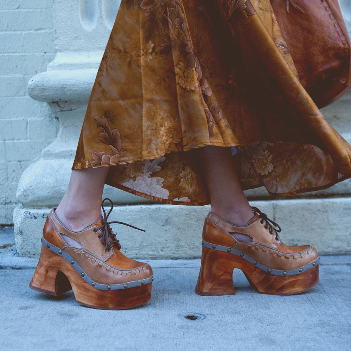 A person wearing the Karaoke long floral skirt and chunky wooden platform shoes by Bed Stu, reminiscent of the 70s era, strides confidently down the sidewalk.