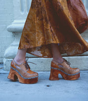 A person wearing the Karaoke long floral skirt and chunky wooden platform shoes by Bed Stu, reminiscent of the 70s era, strides confidently down the sidewalk.
