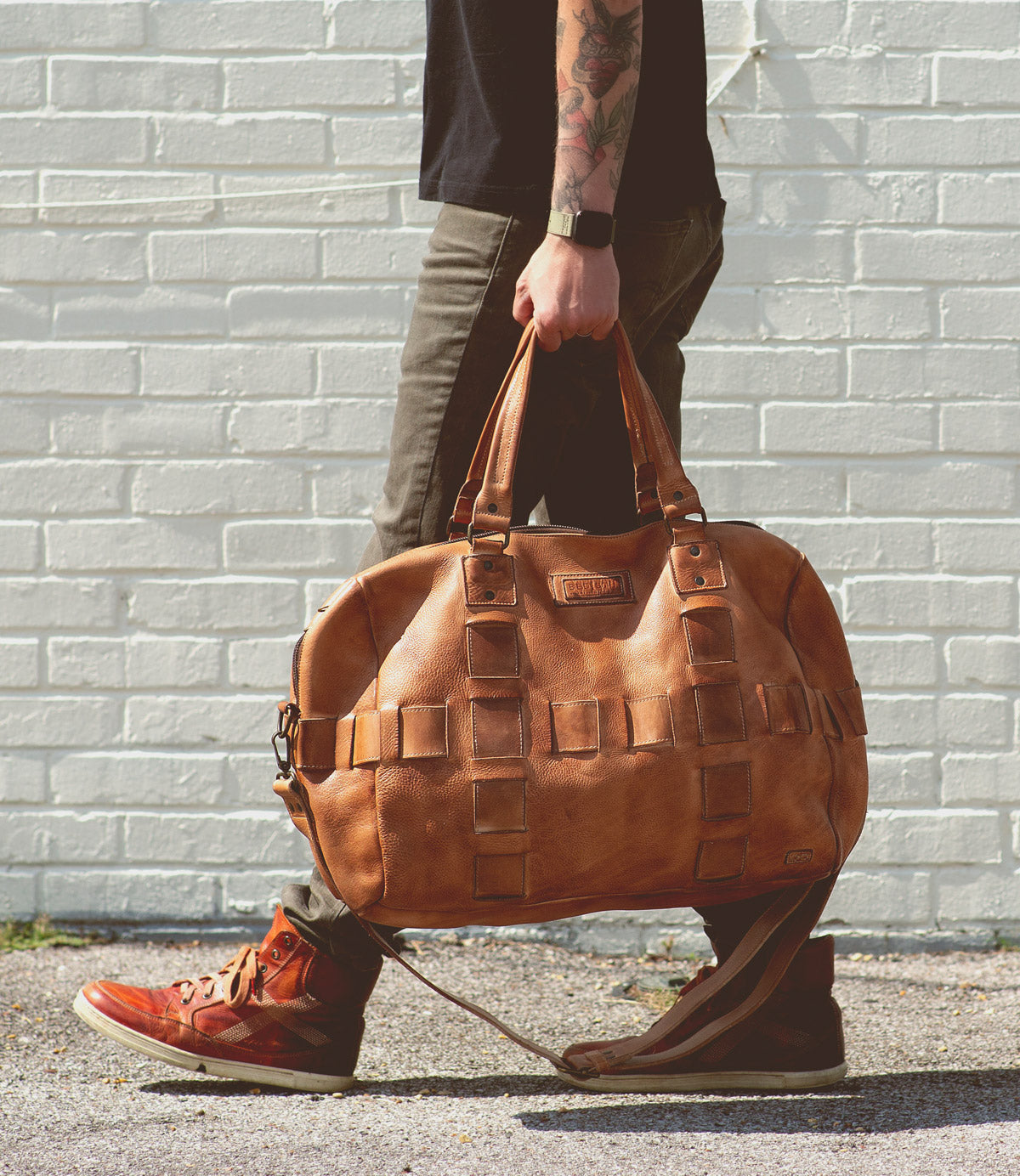 A person with tattooed arms, wearing a black shirt, brown pants, and brown sneakers, is walking while carrying a large handcrafted leather travel duffle bag called the Keg by Bed Stu.