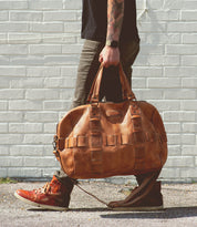 A person with tattooed arms, wearing a black shirt, brown pants, and brown sneakers, is walking while carrying a large handcrafted leather travel duffle bag called the Keg by Bed Stu.