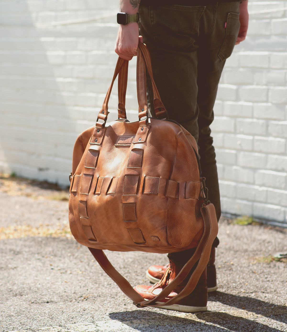 Person holding a large handcrafted Bed Stu Keg leather travel duffle with multiple straps, standing against a brick wall.