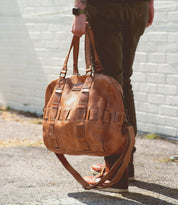Person holding a large handcrafted Bed Stu Keg leather travel duffle with multiple straps, standing against a brick wall.