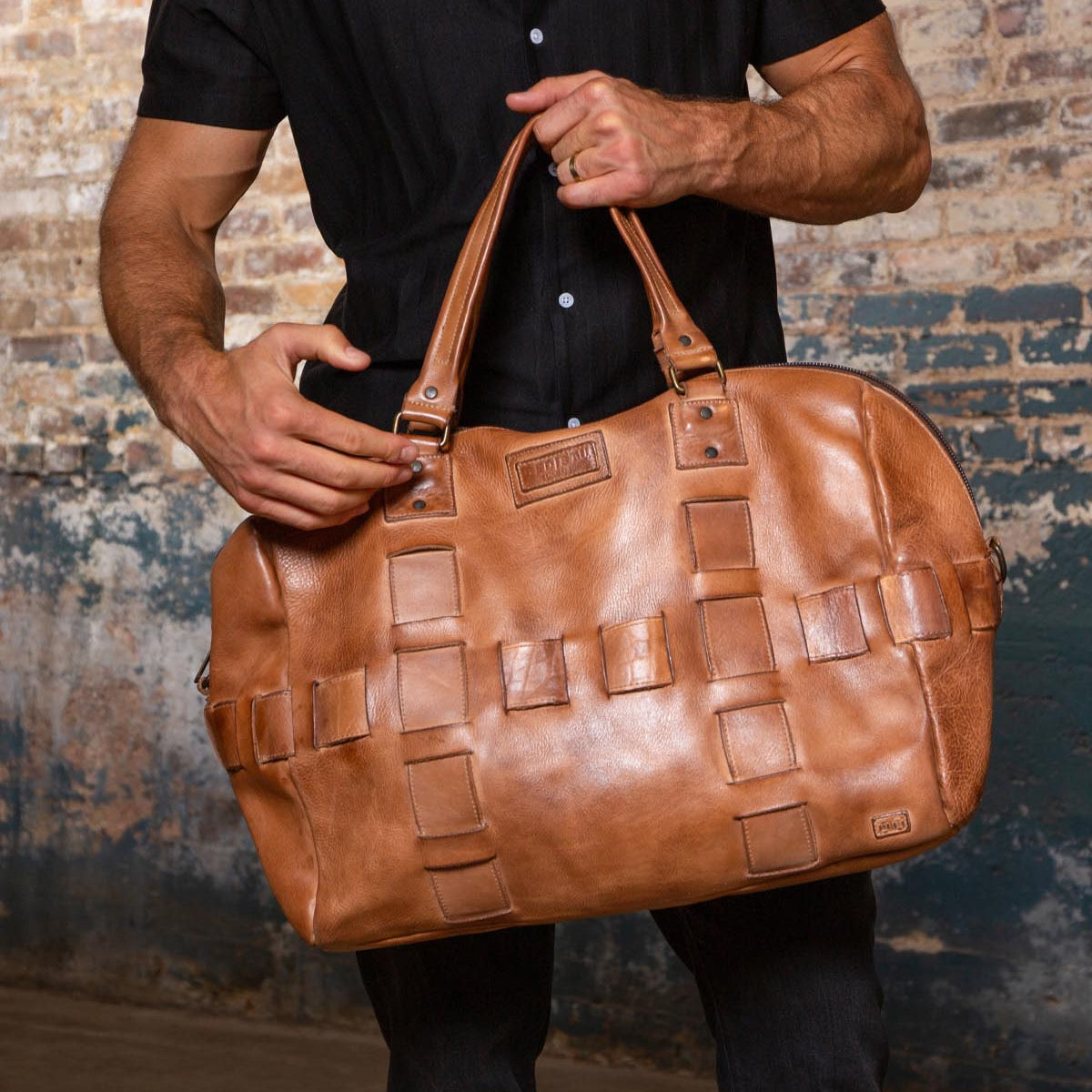 A person in a black shirt holds the Bed Stu Keg, a large tan vegetable-tanned leather duffle with a woven pattern, standing by a rustic brick wall.