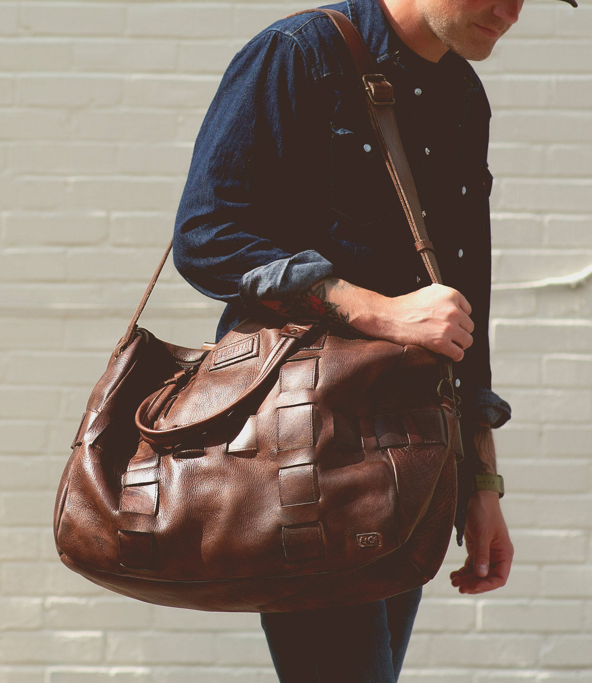 A person in a dark blue shirt carries a large, brown leather Bed Stu Keg shoulder bag made from handcrafted 100% vegetable tanned leather against a white brick wall.