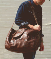 A person in a dark blue shirt carries a large, brown leather Bed Stu Keg shoulder bag made from handcrafted 100% vegetable tanned leather against a white brick wall.