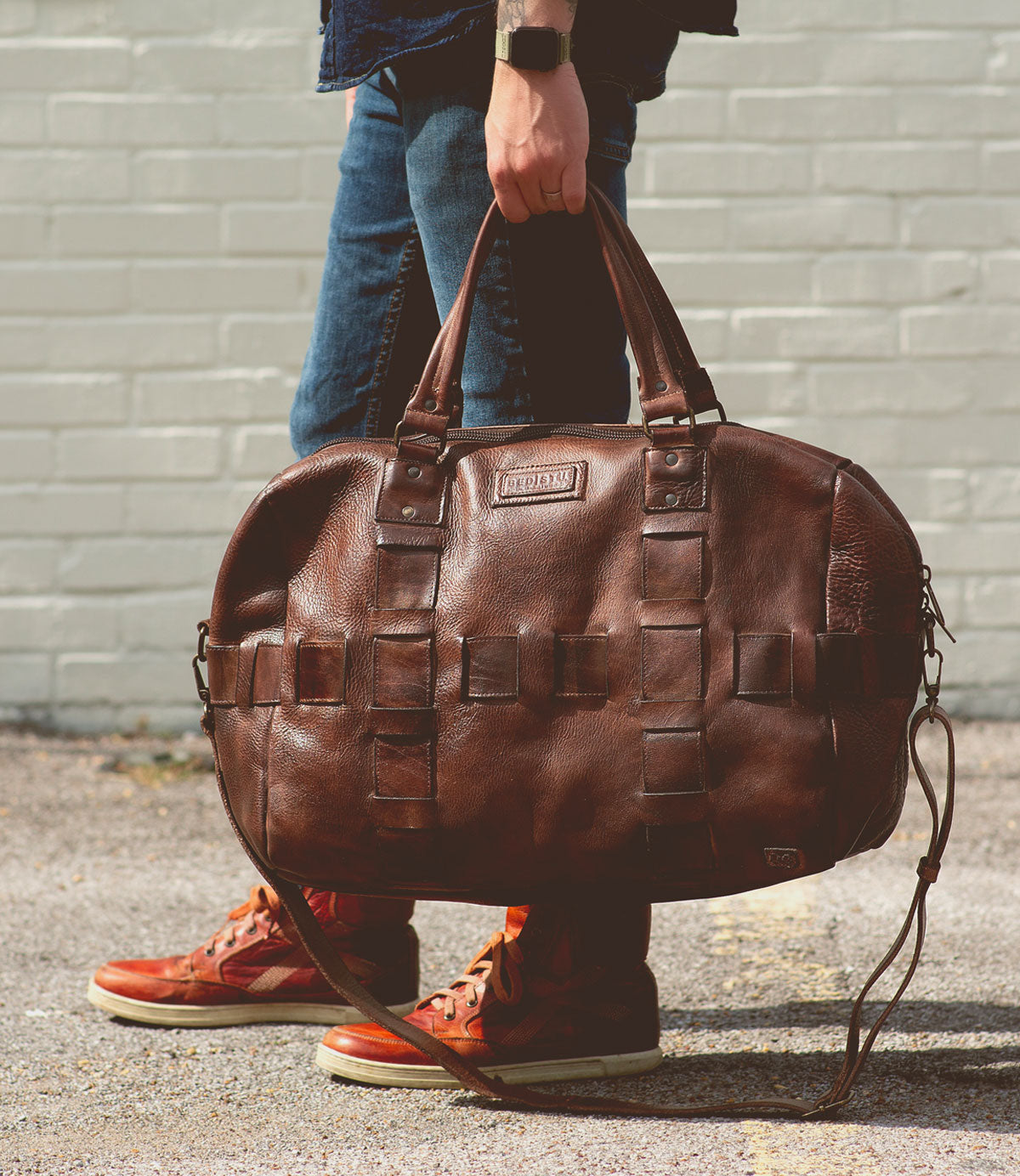 A person holding a large brown Bed Stu Keg duffel bag, made from handcrafted vegetable-tanned leather, wearing blue jeans and red high-top sneakers, stands on a pavement against a light gray brick wall.