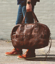A person holding a large brown Bed Stu Keg duffel bag, made from handcrafted vegetable-tanned leather, wearing blue jeans and red high-top sneakers, stands on a pavement against a light gray brick wall.