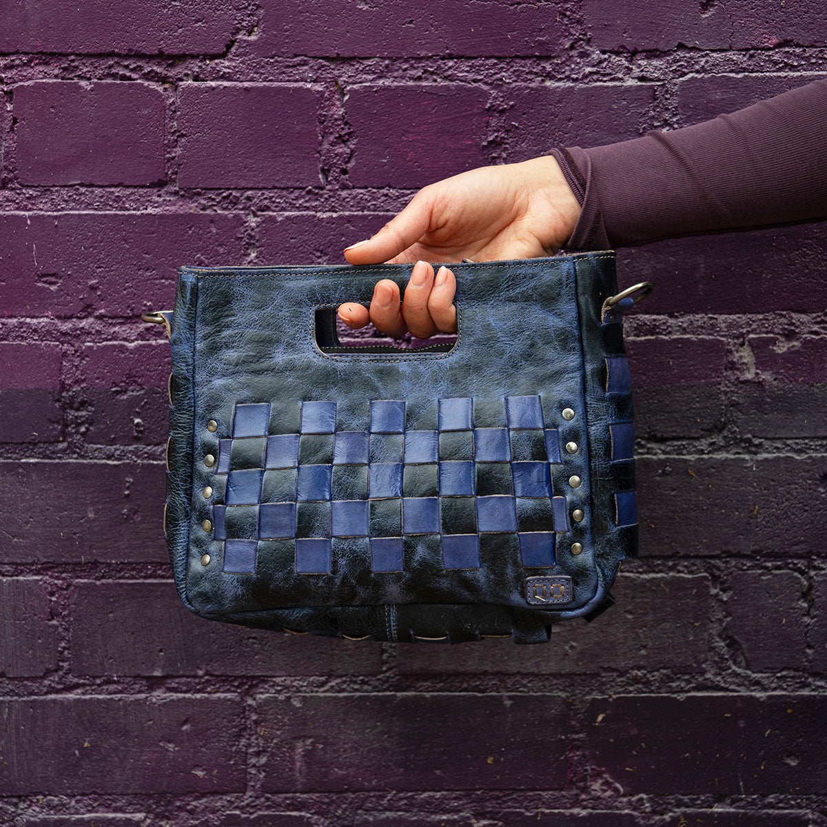 A hand holds the Bed Stu Keiki, a black and blue woven leather clutch with metal studs, against a purple brick wall.