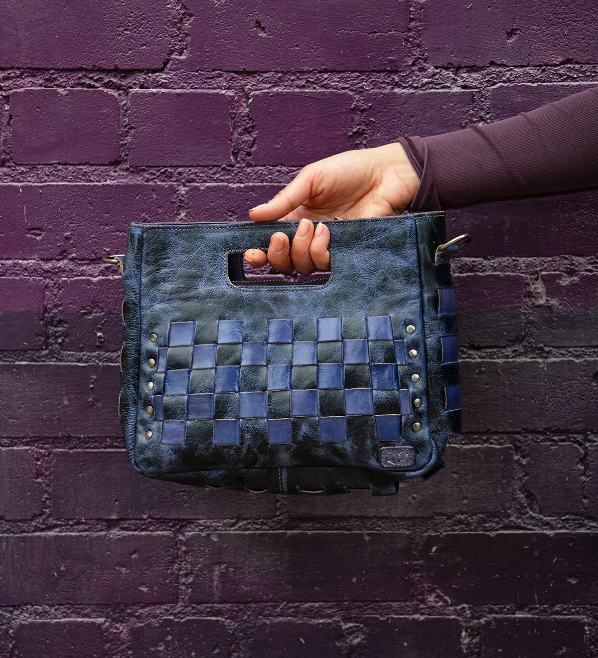 A hand holds the Bed|Stü Keiki clutch—a dark blue woven leather bag with a handle cutout and zip-top closure—in front of a textured purple brick wall.