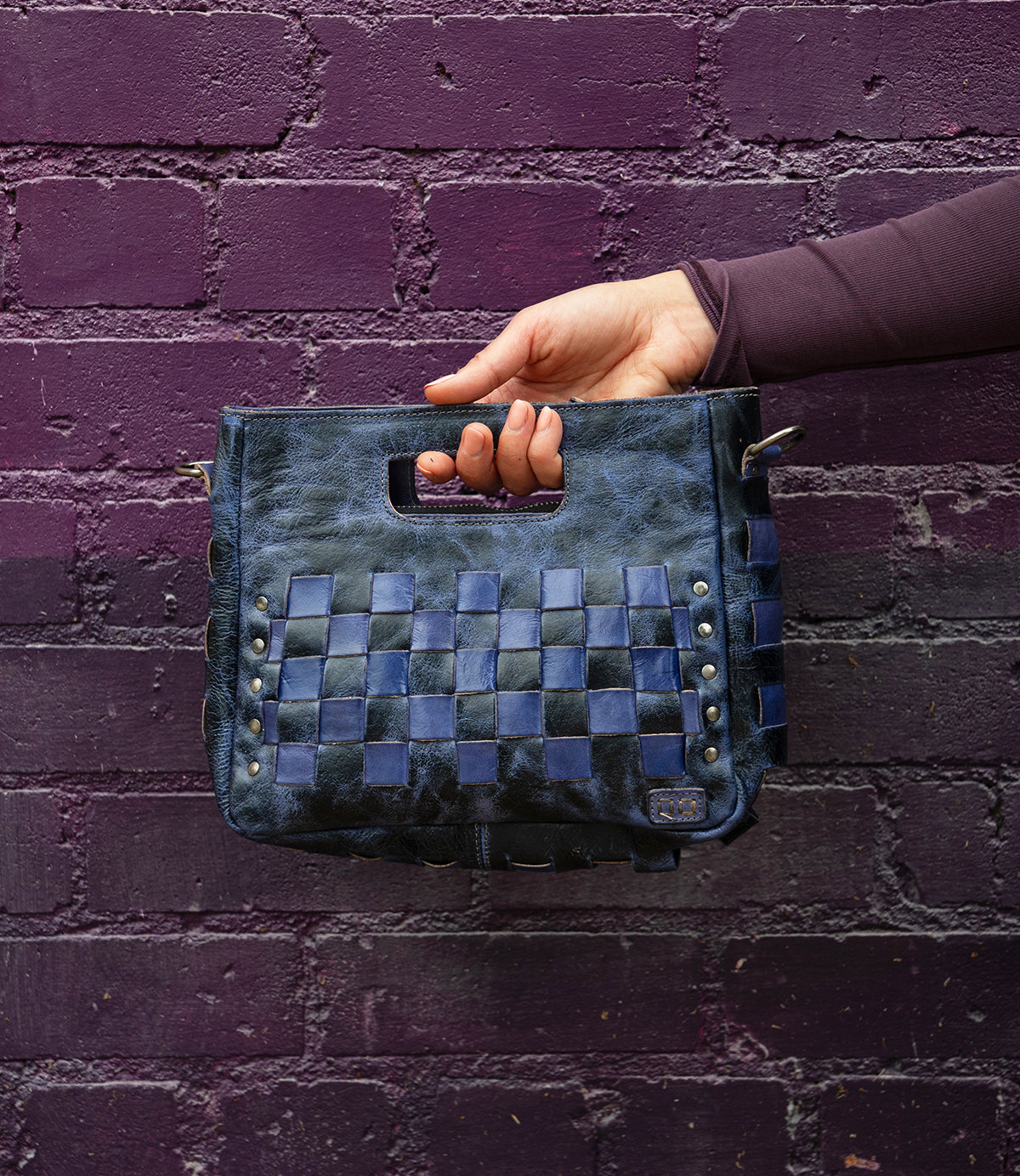A hand holds the Bed Stu Keiki, a black and blue woven leather clutch with metal studs, against a purple brick wall.