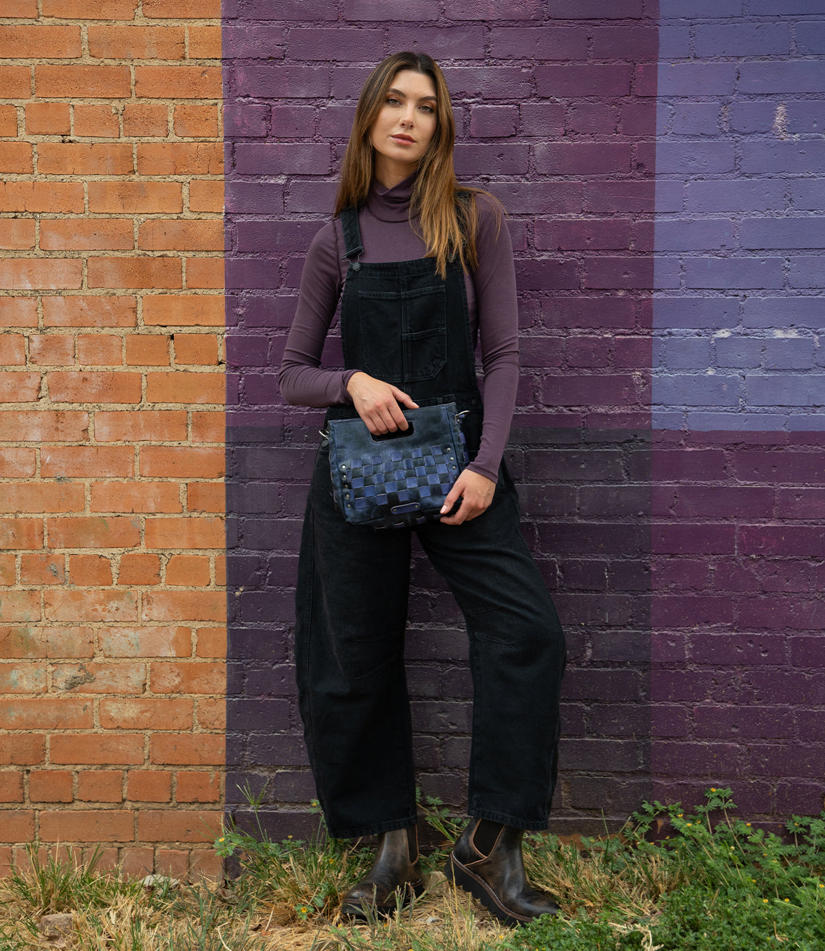 A woman in black overalls and a purple turtleneck stands against an orange and purple brick wall, holding the Bed Stu Keiki blue crossbody bag with woven panels.