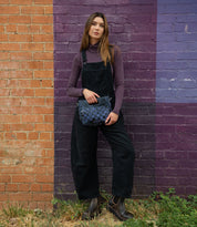 A woman in black overalls and a purple turtleneck stands against an orange and purple brick wall, holding the Bed Stu Keiki blue crossbody bag with woven panels.