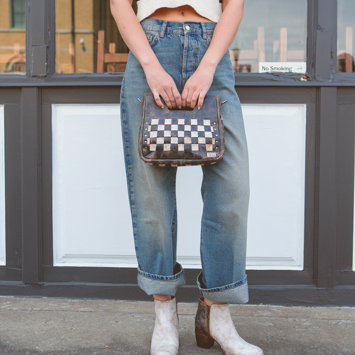 Woman standing in front of a door holding a Bed Stu Keiki checkered crossbody bag with a ziptop closure, wearing cropped white top, blue jeans, and white patterned boots.