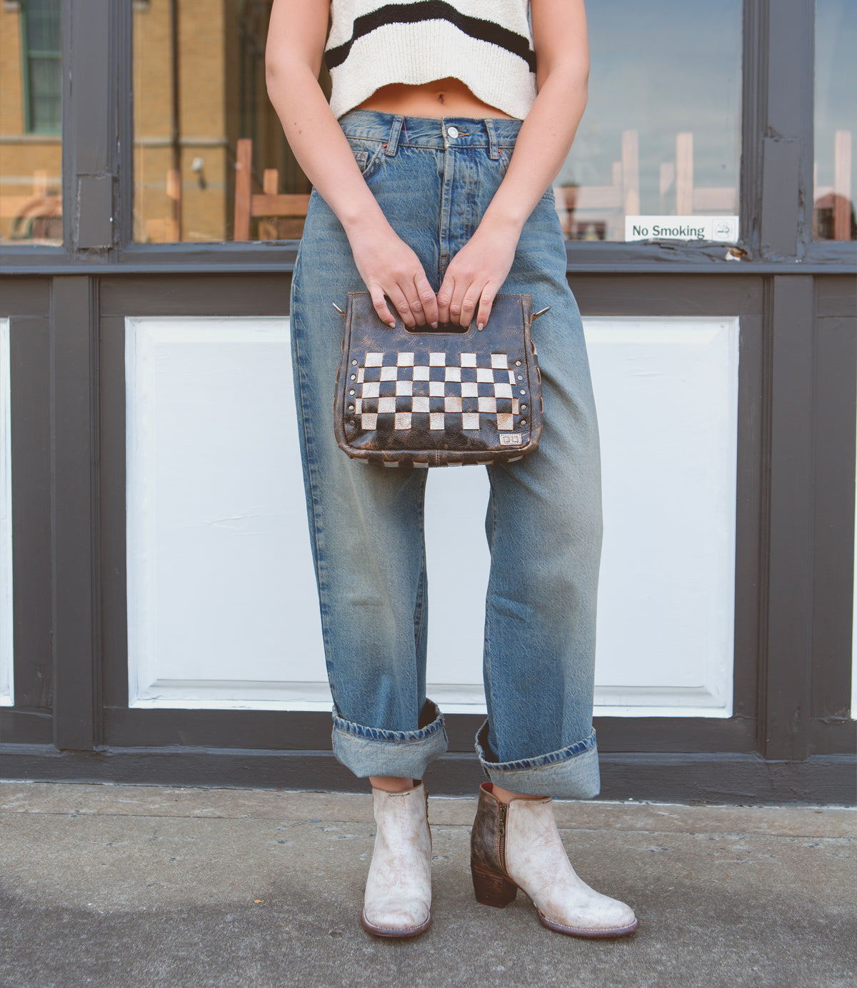 Woman standing in front of a door holding a Bed Stu Keiki checkered crossbody bag with a ziptop closure, wearing cropped white top, blue jeans, and white patterned boots.