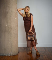 A woman in a brown sheer dress leans against a concrete pillar, holding a woven checkerboard bag as a graphic statement accessory and wearing the Bed|Stü Keiki brown lace-up shoes on a wooden floor.
