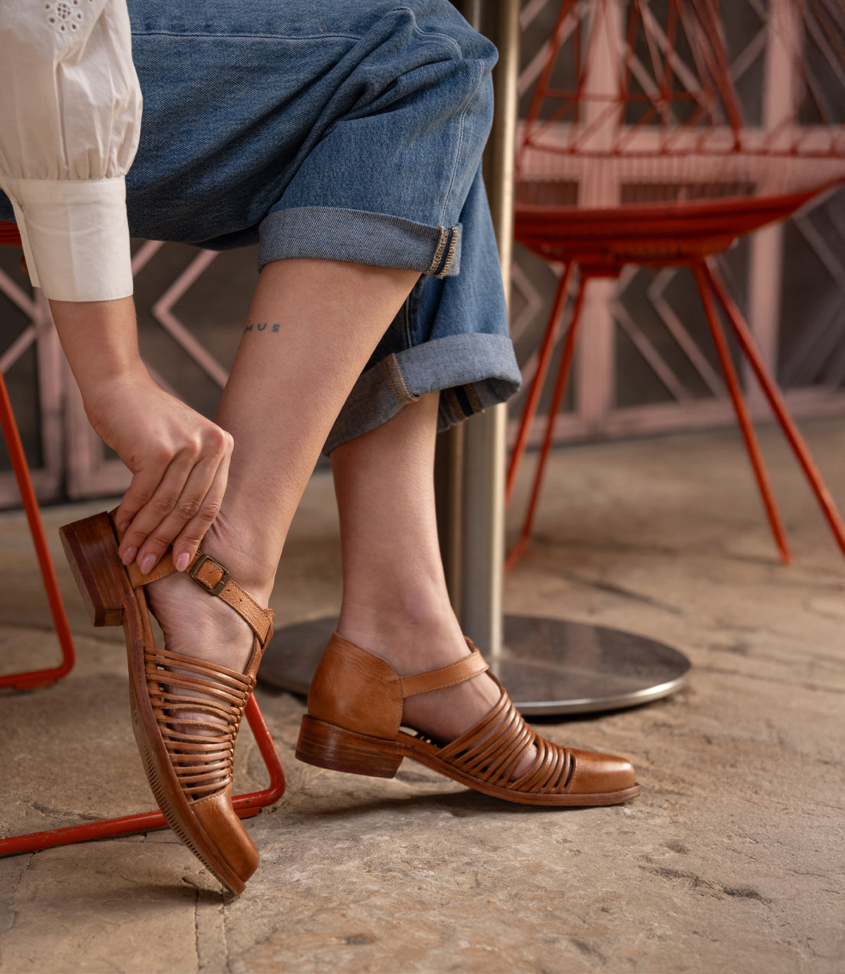 Seated on a red metal chair, a person in cuffed jeans and a white shirt adjusts the Bed Stu Kenna sandal—a brown shoe with woven cutouts and low heel.