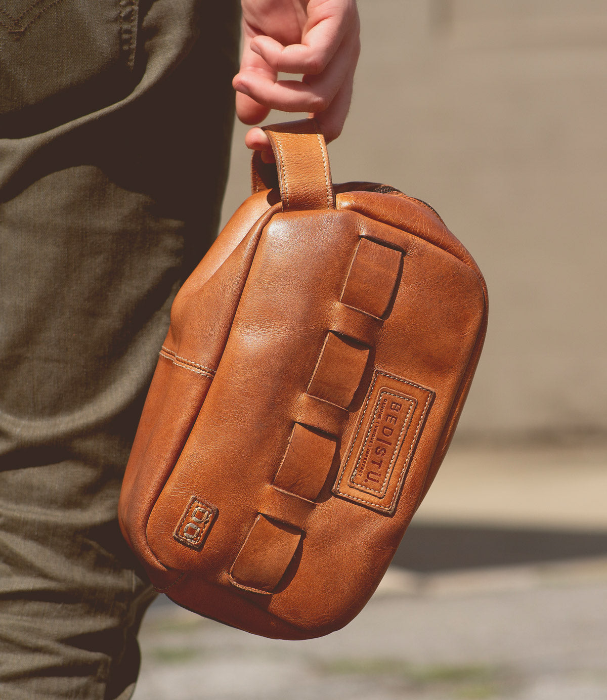 A person carrying a tan leather sling bag featuring a handle and multiple loops, with an embossed Bed Stu logo visible, perfect for travel essentials.