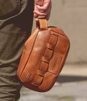 A person carrying a tan leather sling bag featuring a handle and multiple loops, with an embossed Bed Stu logo visible, perfect for travel essentials.