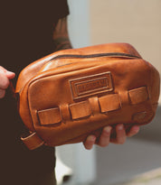 A person holds a brown leather Bed Stu Kip bag with a zippered top and front pockets, perfect for carrying travel essentials.
