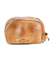 Tan leather dopp kit with a zip closure and a small handle on the side, featuring a metal plate label that reads "Bed Stu." Crafted from vegetable tanned leather, this luxurious Kip bag is perfect for your travel essentials.