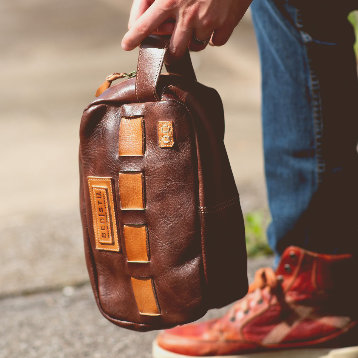 A person holding a brown leather bag with tan accents and visible branding, perfect for carrying travel essentials, while wearing blue jeans and red high-top sneakers.