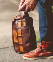 A person holding a brown leather bag with tan accents and visible branding, perfect for carrying travel essentials, while wearing blue jeans and red high-top sneakers.