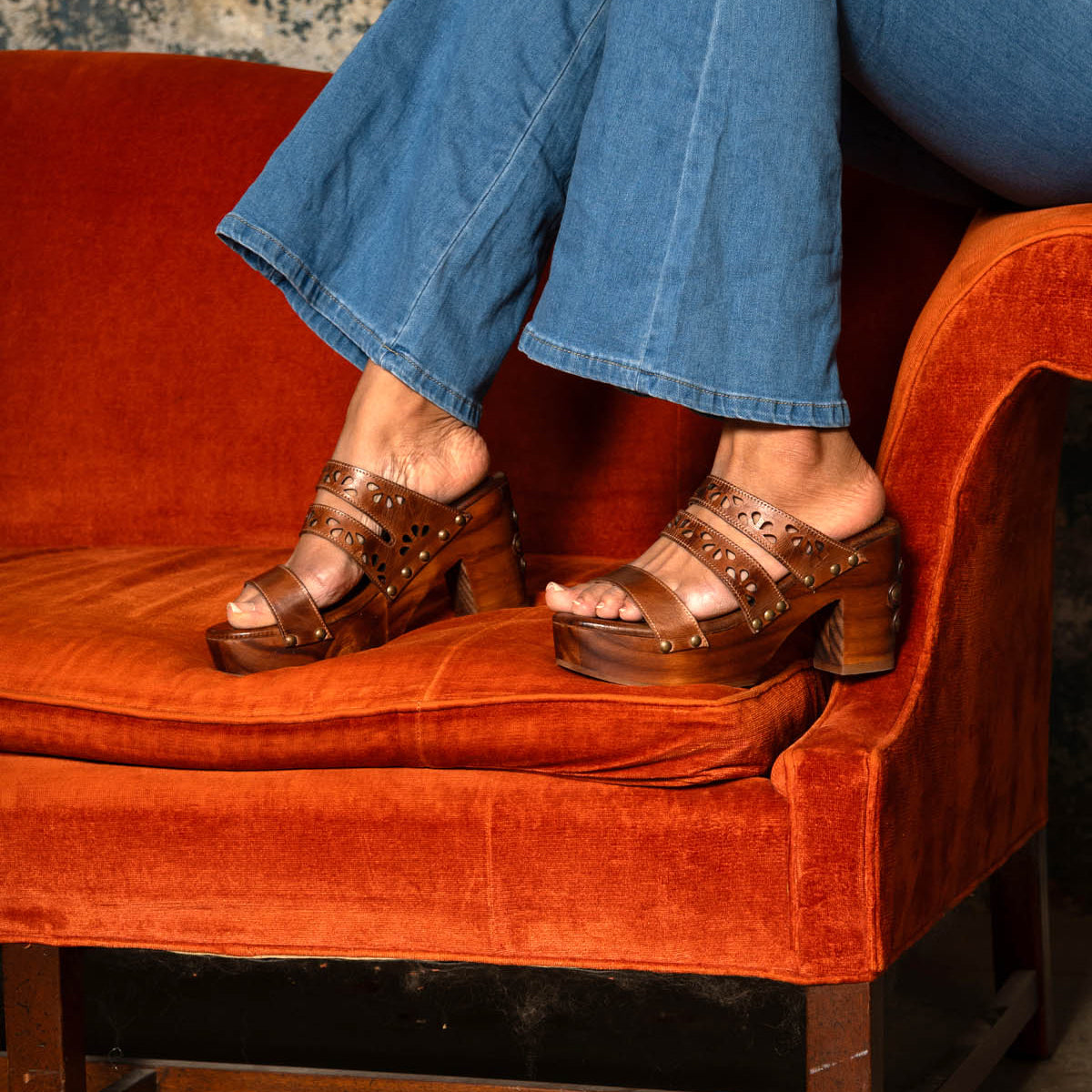 A person wearing Bed Stu's Kreep brown platform sandals with a natural wooden heel sits on an orange velvet couch, legs crossed, paired with blue jeans.
