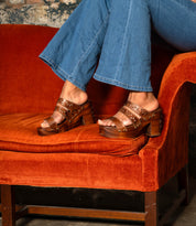A person wearing Bed Stu's Kreep brown platform sandals with a natural wooden heel sits on an orange velvet couch, legs crossed, paired with blue jeans.