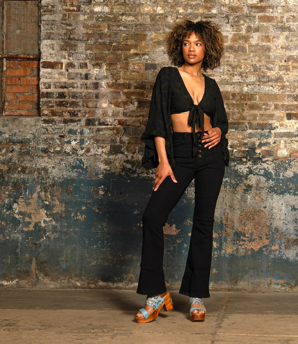 A person stands indoors against a distressed brick wall, wearing black attire and blue Bed Stu Kreep platform sandals with wooden 70's-inspired heels, exuding whimsical confidence.