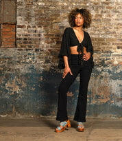 A person stands indoors against a distressed brick wall, wearing black attire and blue Bed Stu Kreep platform sandals with wooden 70's-inspired heels, exuding whimsical confidence.