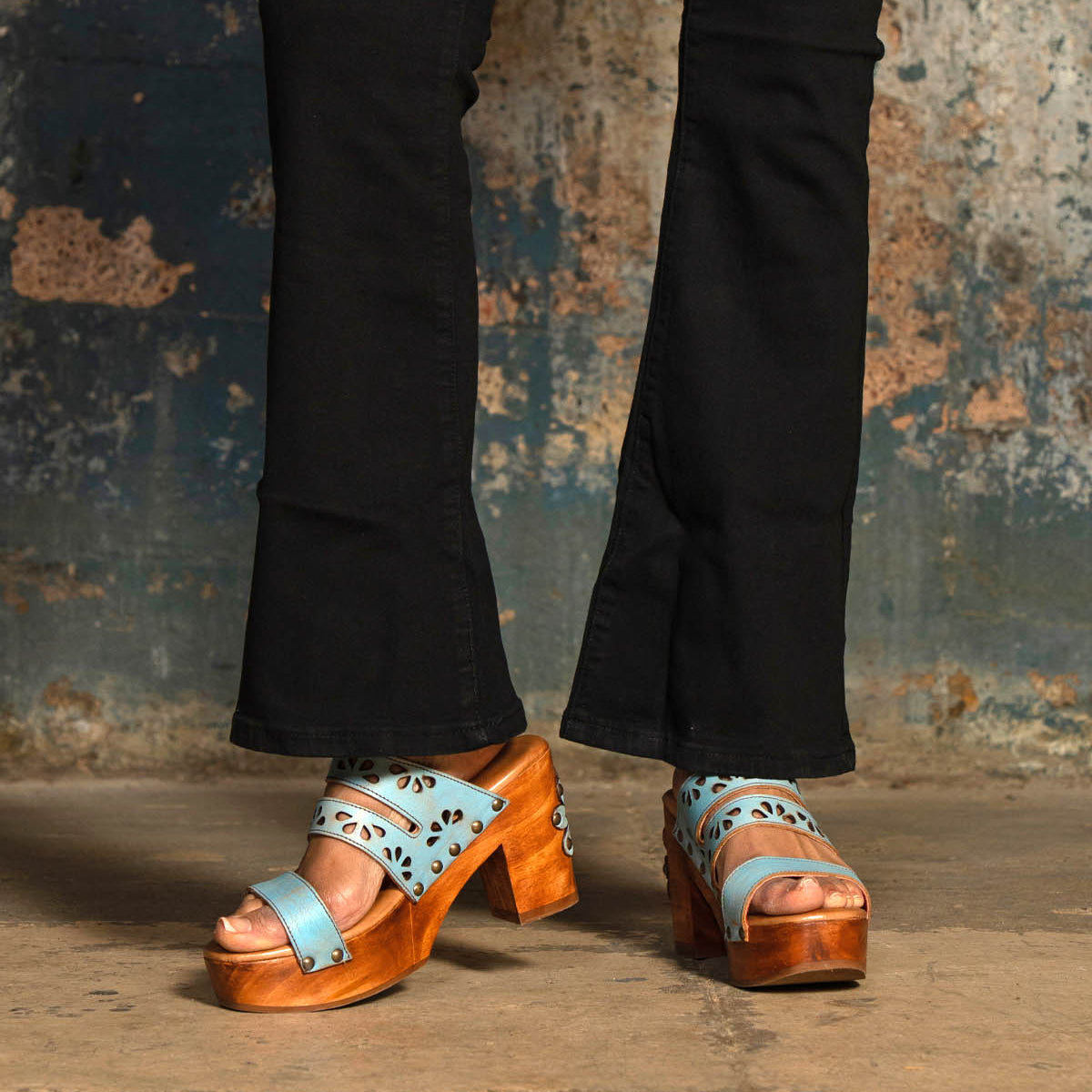A person wearing Bed Stu's Kreep black pants and blue leather platform sandals with floral cutouts and a chunky wooden heel stands on a concrete floor against a textured wall.