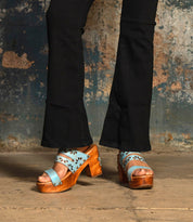 A person wearing Bed Stu's Kreep black pants and blue leather platform sandals with floral cutouts and a chunky wooden heel stands on a concrete floor against a textured wall.