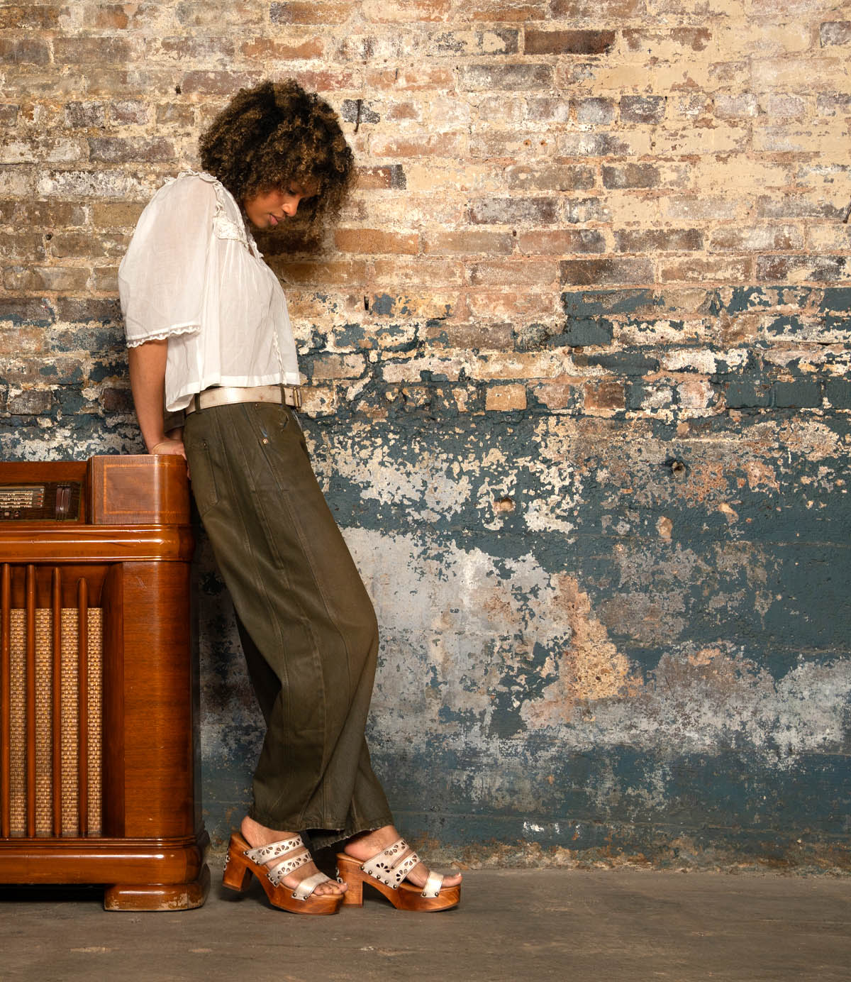 Wearing Bed Stu's Kreep—70's-inspired heels with natural wooden heels—a person in a white blouse and green pants leans on a vintage wooden radio before a distressed brick wall.