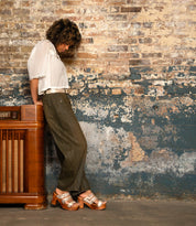 Wearing Bed Stu's Kreep—70's-inspired heels with natural wooden heels—a person in a white blouse and green pants leans on a vintage wooden radio before a distressed brick wall.