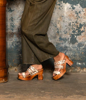 Wearing Bed Stu's Kreep olive green pants and 70s-inspired studded heels, a person stands confidently on a concrete floor beside a wooden table against a textured blue-gray wall.