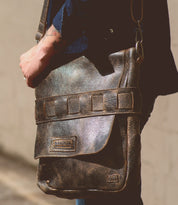 A person is carrying a weathered messenger bag made from vegetable tanned leather. The brown leather shoulder bag, named Kure, by Bed Stu features a large flap and a clasp, along with a visible brand label. It also has an artisanal touch with its handwoven exterior strap.