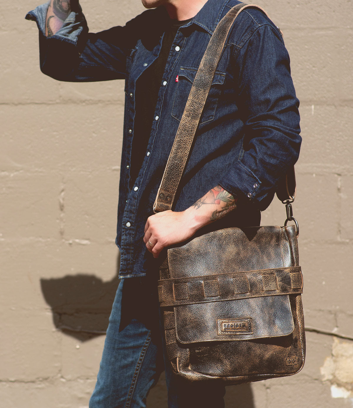 A person wearing a denim shirt and jeans holds a Bed Stu Kure with a handwoven exterior strap over their shoulder, standing against a beige concrete wall.