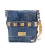 A blue leather crossbody bag with a tan strap and square tan accents on the front flap. The brand logo "Bed Stu" is displayed on a patch at the front. Crafted from vegetable tanned leather, this handmade Kure messenger bag boasts an organic cotton-lined interior.