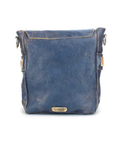 A square, dark blue handmade leather messenger bag with a metallic logo plate on the front, featuring side buckles, a flap closure, and an organic cotton-lined interior called Kure by Bed Stu.