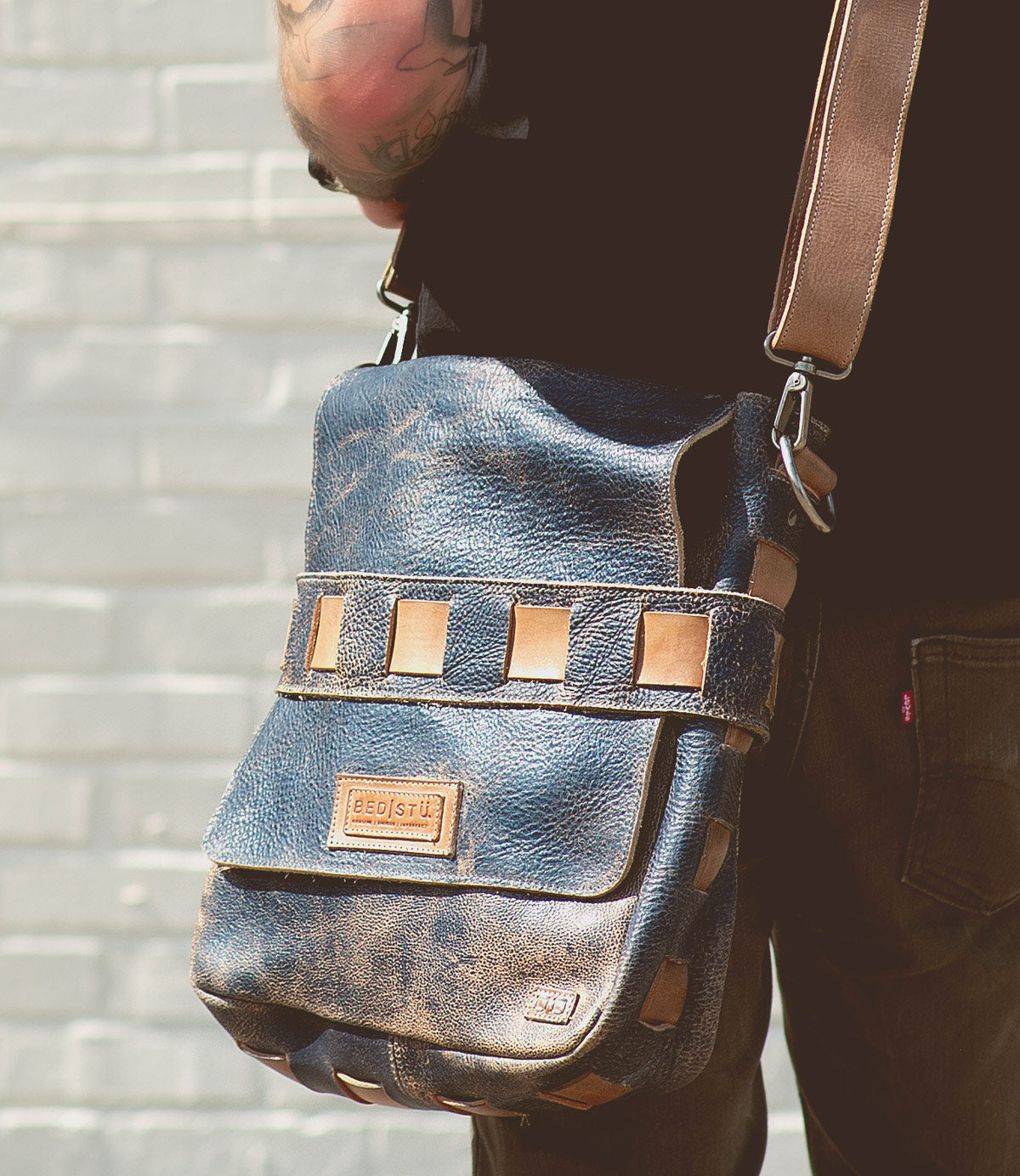 A person with a tattooed arm holds the Kure crossbody bag by Bed Stu, with beige accents, featuring a handwoven exterior strap and a text label on the front, against a light brick wall.