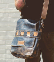 A person with a tattooed arm holds the Kure crossbody bag by Bed Stu, with beige accents, featuring a handwoven exterior strap and a text label on the front, against a light brick wall.