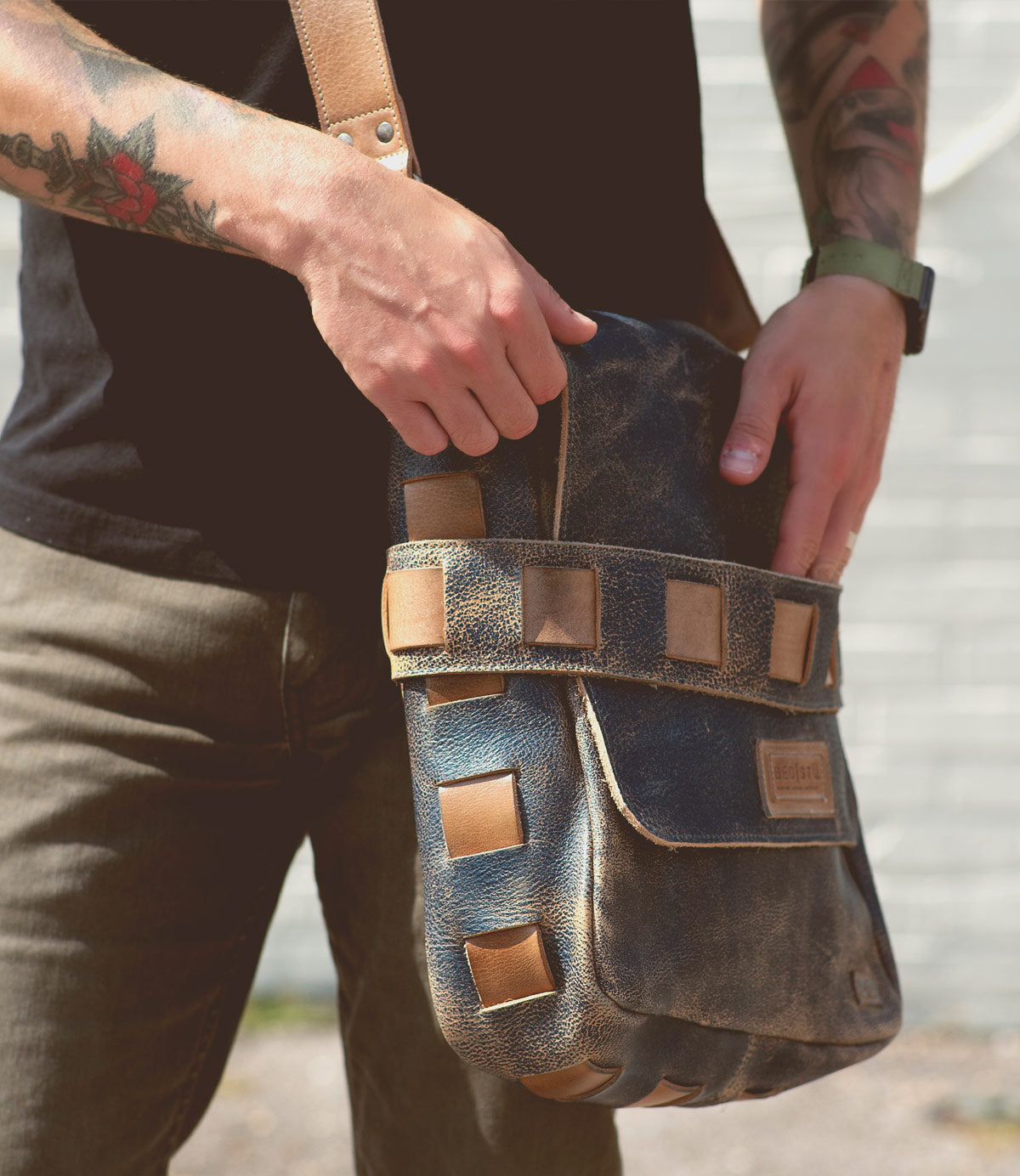 A person with tattoos and wearing a watch carries a worn leather shoulder bag, crafted from vegetable tanned leather. They hold the handwoven exterior strap of the Kure by Bed Stu with one hand and adjust the bag with the other, against a blurred background.