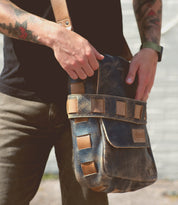 A person with tattoos and wearing a watch carries a worn leather shoulder bag, crafted from vegetable tanned leather. They hold the handwoven exterior strap of the Kure by Bed Stu with one hand and adjust the bag with the other, against a blurred background.