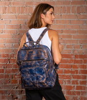 A woman in a white tank top and black pants stands before a red brick wall, wearing the Bed Stu Lafe—a large, distressed blue leather backpack that doubles as a stylish work bag.