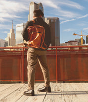 Wearing Bed Stu's Barton leather high-top sneakers with breathable insoles, a person with a brown backpack stands on a wooden platform. They face the city skyscrapers and a crane in the distance under a blue sky, ready for adventure at every comfortable step.