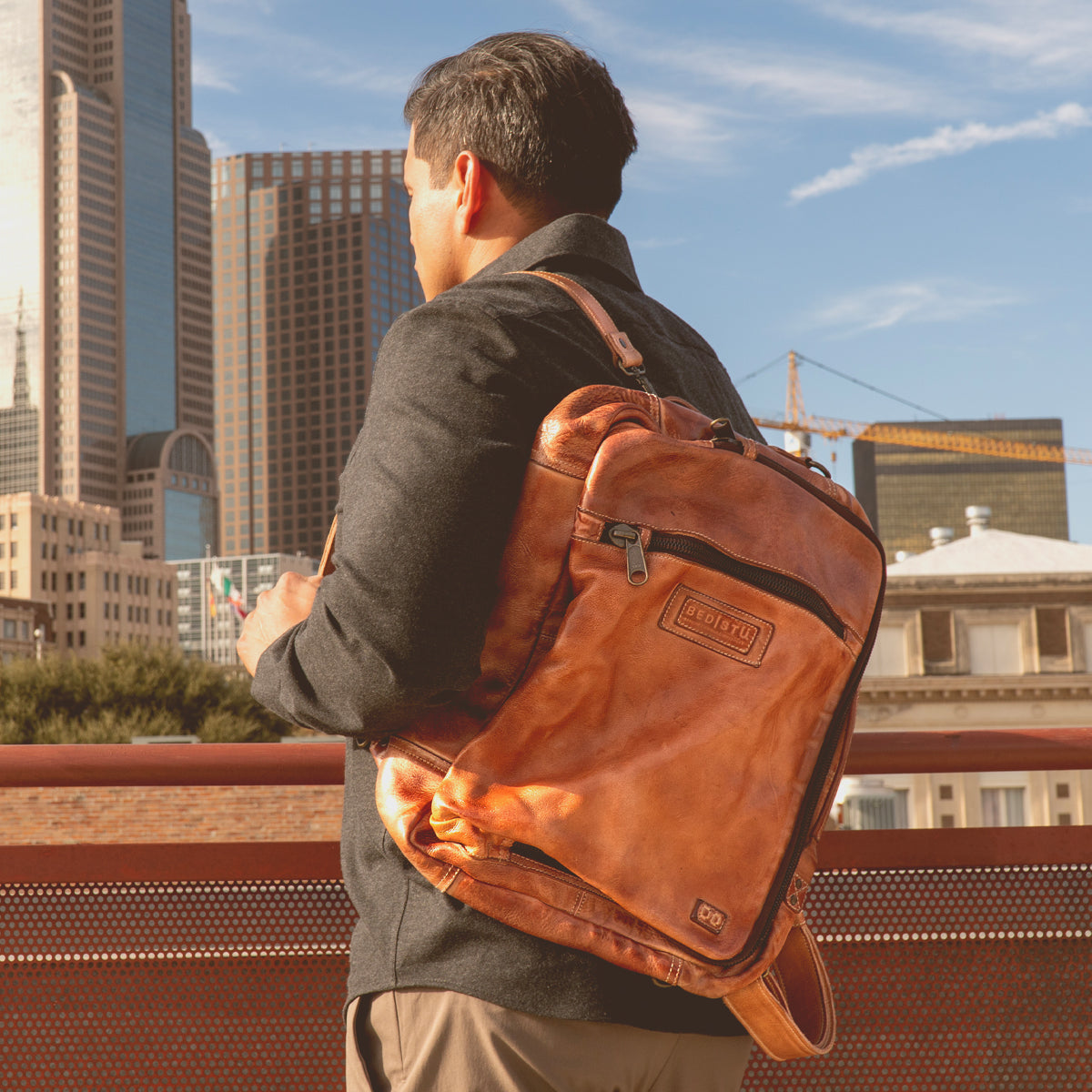 A man with a Barton leather backpack and Bed Stu stylish leather high-top sneakers stands on a rooftop, overlooking a cityscape with tall buildings under a clear sky.
