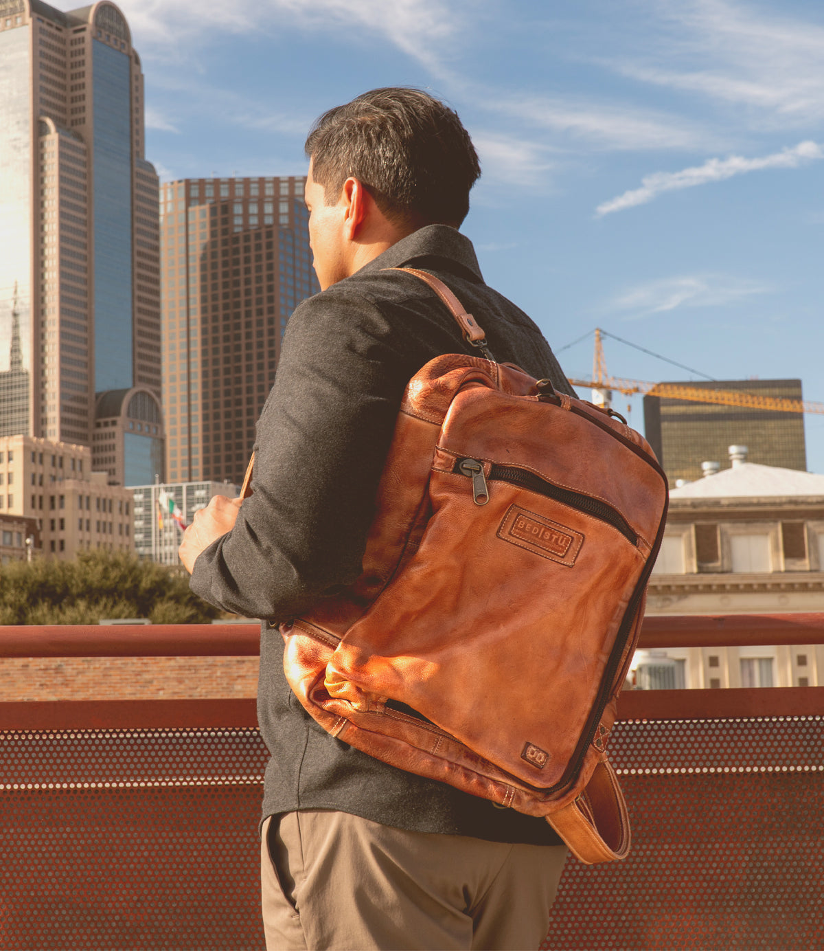 A man with a Barton leather backpack and Bed Stu stylish leather high-top sneakers stands on a rooftop, overlooking a cityscape with tall buildings under a clear sky.