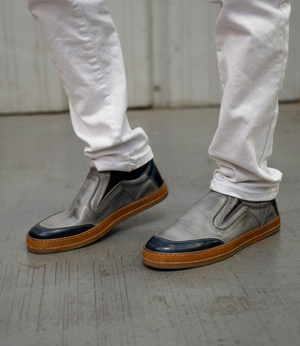 A person wearing white pants and Bed|Stü Lanius slip-on shoes with gray uppers, black accents, and brown soles stands on a concrete floor. The Lanius shoes feature subtle color blocking details, creating a fresh yet timeless silhouette.