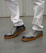 A person wearing white pants and Bed|Stü Lanius slip-on shoes with gray uppers, black accents, and brown soles stands on a concrete floor. The Lanius shoes feature subtle color blocking details, creating a fresh yet timeless silhouette.