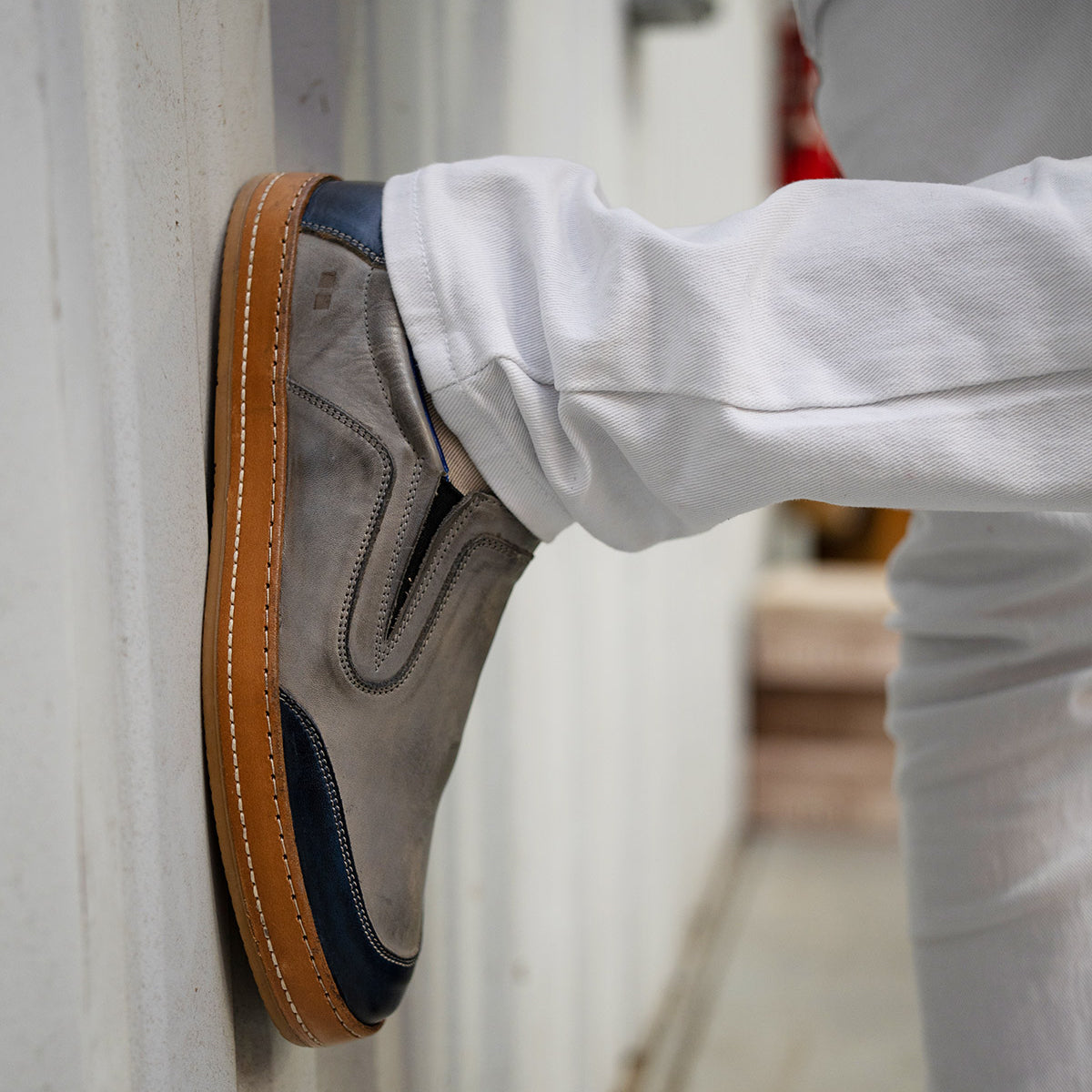 Wearing crisp white pants, the person showcases the timeless silhouette of Bed|Stü’s Lanius slip-on shoes—crafted in gray suede with navy accents and gum soles—as they rest their foot against a wall.